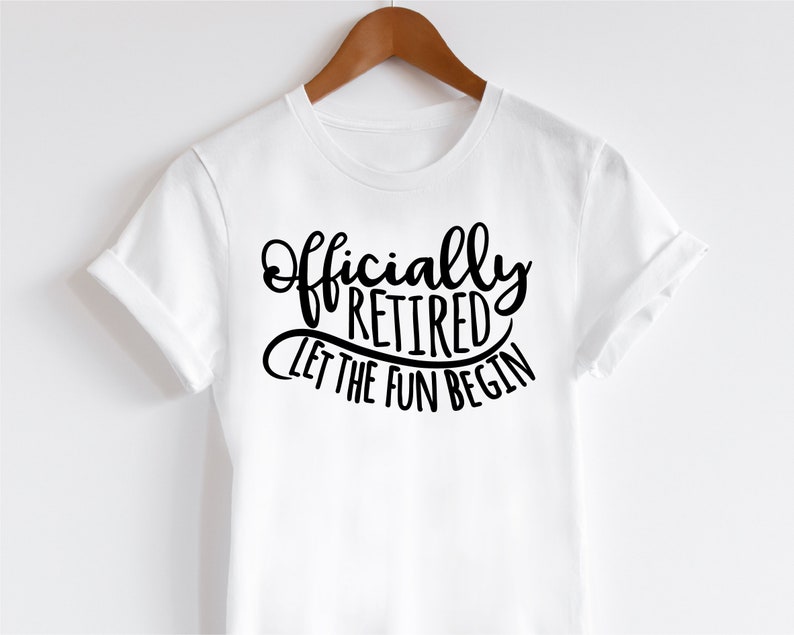 May include: A white t-shirt with the words "Officially Retired Let The Fun Begin" in black script. The shirt is hanging on a wooden hanger.