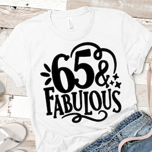Puede incluir: Camiseta blanca con la frase "65 & Fabulous" en negro. El diseño incluye el número 65, el símbolo "&" y la palabra "Fabulous" con elementos decorativos. La camiseta se muestra con unos vaqueros azules.