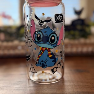 May include: A clear glass jar with a pink lid, decorated with a blue cartoon character wearing glasses and a scarf. The jar also features various Harry Potter-themed graphics, including a golden snitch and a Hogwarts letter.