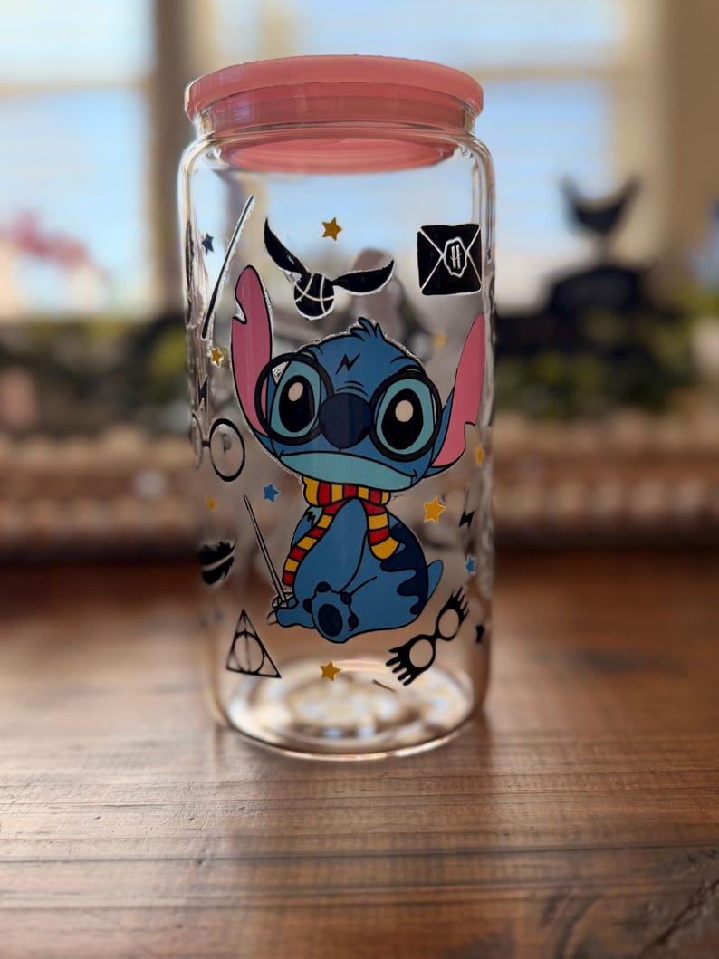 May include: A clear glass jar with a pink lid, decorated with a blue cartoon character wearing glasses and a scarf. The jar also features various Harry Potter-themed graphics, including a golden snitch and a Hogwarts letter.