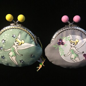 May include: Two coin purses with a metal clasp and decorative yellow and pink ball closures. Each purse features a cartoon image of Tinkerbell. One purse is light green with stars, the other is gray.