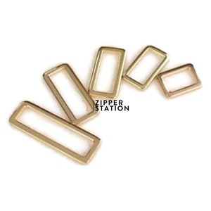 May include: Five gold-coloured metal rectangular rings of different sizes. The rings are arranged in a semi-circle shape. The rings are smooth and have a shiny finish.