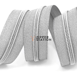 May include: A close-up of a silver zipper tape with a herringbone pattern. The zipper tape is coiled and the zipper teeth are visible.