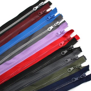 May include: Assortment of zipper closures in various colours, including red, blue, purple, black, grey, and green. The zippers are arranged in a fan shape, with the zipper teeth facing up.