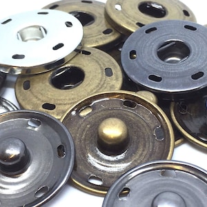 May include: Assortment of metal snap buttons in silver, brass, and black finishes. The buttons are circular with a raised centre and a decorative ring around the edge.