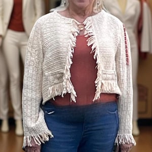 May include: Cream-colored, textured jacket with fringe detailing. The open-front cardigan features a round neckline and long sleeves. The jacket is paired with a rust-colored top and blue jeans. The overall style is casual and bohemian.