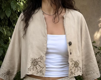 May include: A beige, cropped jacket with floral embroidery on the sleeves and front. The jacket has a single button closure and is worn over a white tube top. The sleeves are three-quarter length.