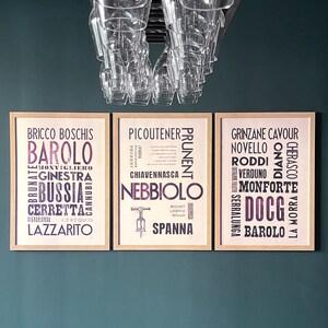 May include: Three framed posters with a wine theme. The posters feature the names of different Italian wines, including Barolo, Nebbiolo, and Barbera. The posters are hung on a dark green wall.