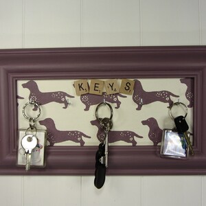 May include: A purple picture frame with a white background featuring a dachshund pattern and the word "KEYS" spelled out with letter tiles.  There are three keychains hanging from hooks on the frame.
