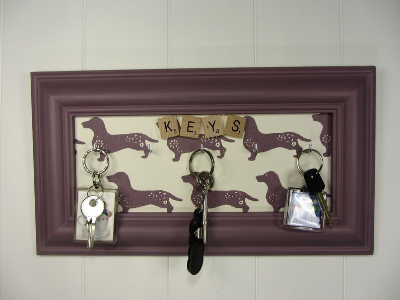 May include: A purple picture frame with a white background featuring a dachshund pattern and the word "KEYS" spelled out with letter tiles.  There are three keychains hanging from hooks on the frame.