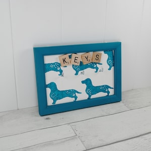 May include: A teal blue framed key holder with a white background featuring a repeating pattern of blue dachshunds. The word "KEYS" is spelled out using wooden letter tiles.