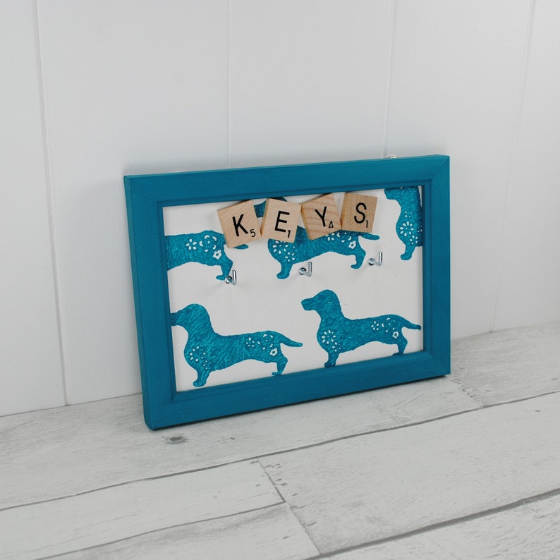 May include: A teal blue framed key holder with a white background featuring a repeating pattern of blue dachshunds. The word "KEYS" is spelled out using wooden letter tiles.