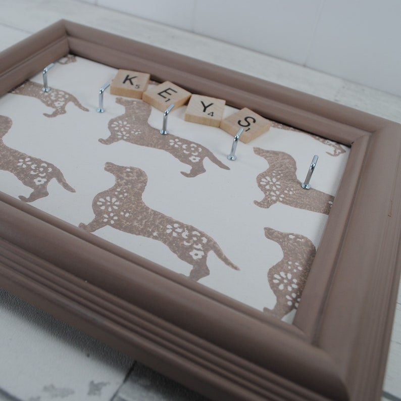 May include: A brown wooden frame with a white background and a pattern of brown dachshunds. The frame has five hooks and the word "KEYS" spelled out with letter tiles.