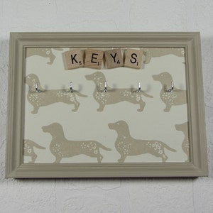 May include: A beige key holder with a white background featuring a repeating pattern of dachshund silhouettes. The key holder has five hooks and the word "KEYS" spelled out with letter tiles.