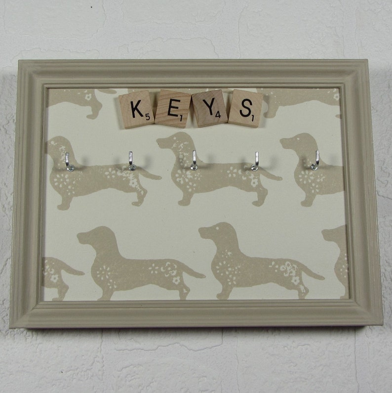 May include: A beige key holder with a white background featuring a repeating pattern of dachshund silhouettes. The key holder has five hooks and the word "KEYS" spelled out with letter tiles.