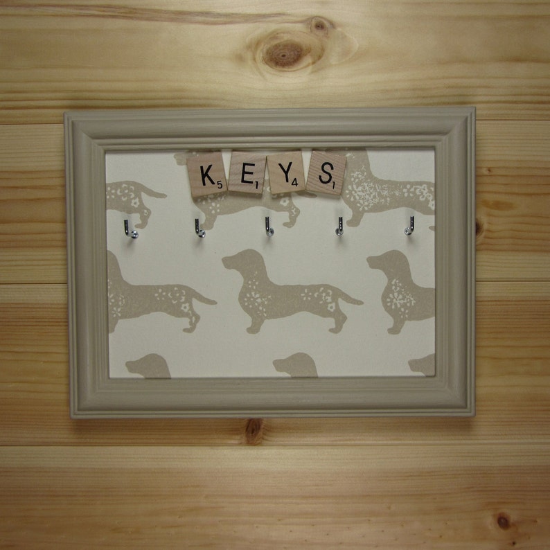 May include: A gray framed key holder with a beige background featuring a repeating pattern of dachshund dogs. The word "KEYS" is spelled out using letter tiles.