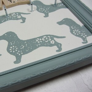May include: A light blue picture frame with a white background and a pattern of gray dachshunds with white floral designs. The frame has a decorative edge.