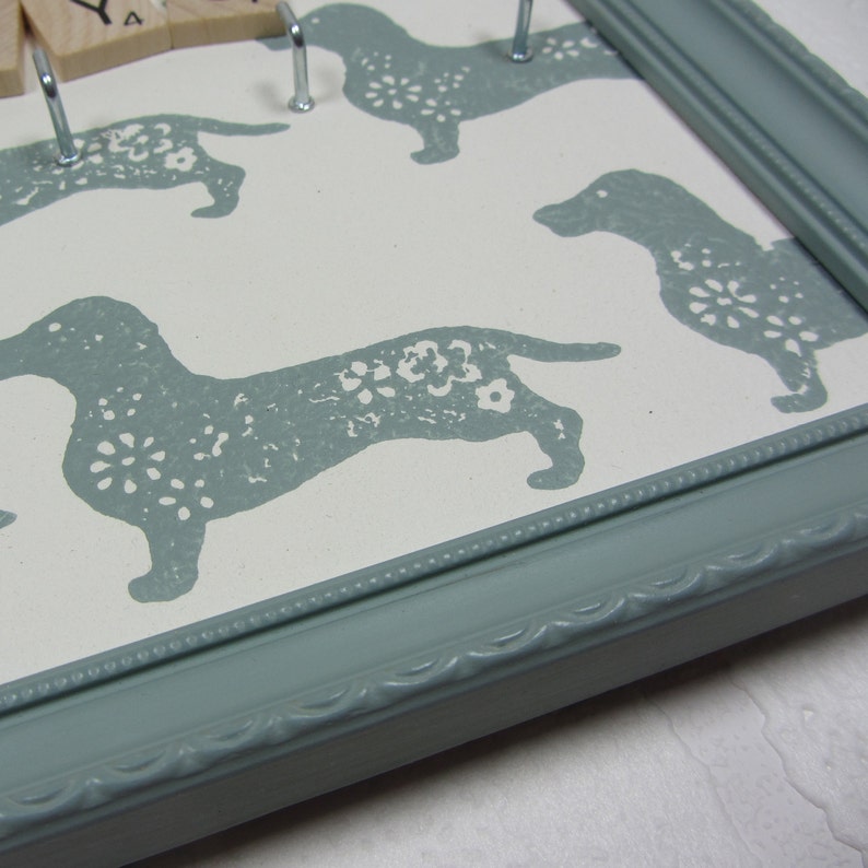 May include: A light blue picture frame with a white background and a pattern of gray dachshunds with white floral designs. The frame has a decorative edge.