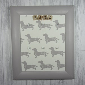 May include: A gray framed key holder with a white background featuring a repeating pattern of gray dachshunds. The word "KEYS" is spelled out using wooden letter tiles.