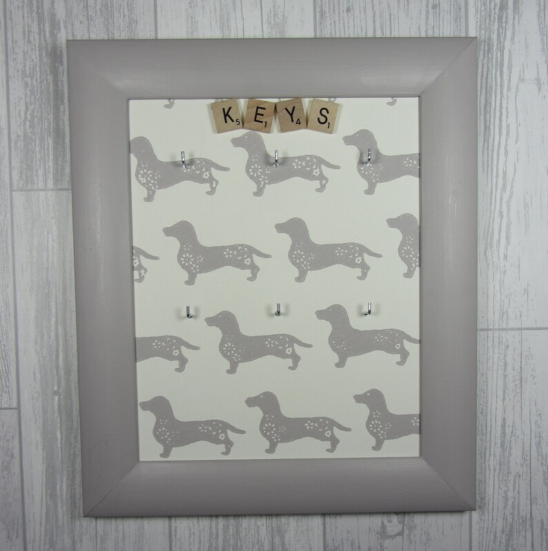 May include: A gray framed key holder with a white background featuring a repeating pattern of gray dachshunds. The word "KEYS" is spelled out using wooden letter tiles.