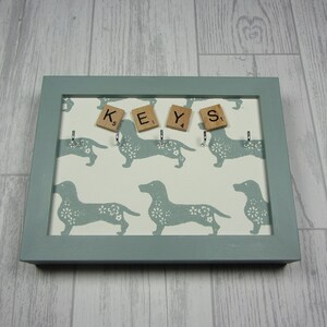 May include: A light blue key holder with a white background featuring a repeating pattern of dachshund silhouettes. The key holder has five hooks and the word "KEYS" spelled out with wooden letter tiles.