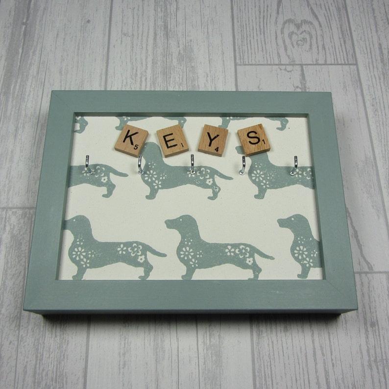 May include: A light blue key holder with a white background featuring a repeating pattern of dachshund silhouettes. The key holder has five hooks and the word "KEYS" spelled out with wooden letter tiles.