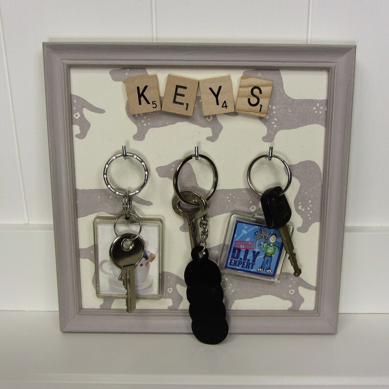 May include: A gray framed key holder with a white background featuring a repeating pattern of black dachshund silhouettes. The holder has the word "KEYS" spelled out with letter tiles. There are three key rings with keys and a small keychain with a blue and white graphic of a person with the text "DIY EXPERT".