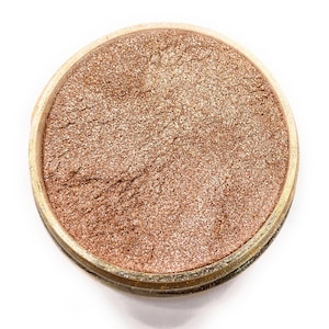 May include: A round container filled with a fine, shimmery, light brown powder. The powder has a subtle golden sheen.