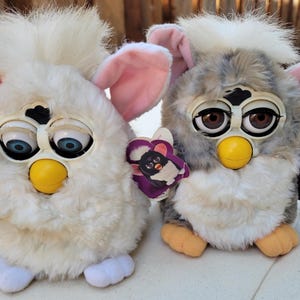 May include: Two plush electronic toys with soft fur, large plastic eyes, and yellow beaks. The left toy is solid white with blue eyes and white feet. The right toy is grey and white with brown eyes, orange feet, and a purple tag attached.