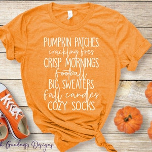 May include: Orange t-shirt with white text that reads "Pumpkin Patches, crackling fires, CRISP MORNINGS, Football, BIG SWEATERS, fall candles, COZY SOCKS". The shirt is tied in a knot and is laying on a wooden surface with two pairs of orange converse shoes and three small orange pumpkins.