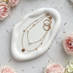 May include: A gold necklace with a pearl pendant and a gold coin pendant, along with two gold rings, are displayed on a white, organically shaped tray. The tray is surrounded by pink roses, white flowers, and a white satin cloth.