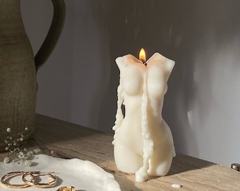 Puede incluir: Una vela de color crema con forma de torso femenino, con una mecha encendida. La vela se está derritiendo, con cera goteando. Joyas doradas y un plato decorativo en primer plano. La vela está sobre una superficie clara.