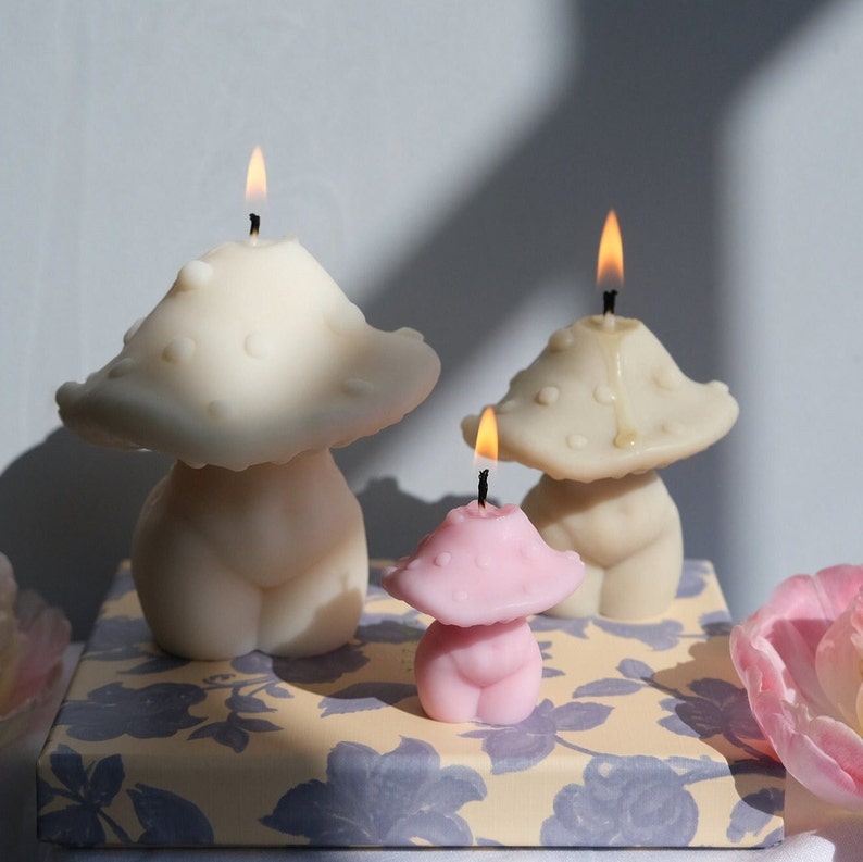 May include: Three mushroom-shaped candles, one pink, one beige and one white, with flames lit. The candles are on a floral patterned surface.