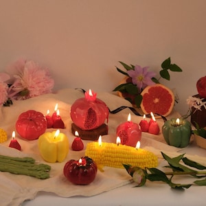 Op de afbeelding: Een verzameling kaarsen in de vorm van groenten en fruit, waaronder een rode granaatappel, gele en groene paprika, een ma&iuml;skolf en kleine aardbeien. Verschillende kaarsen branden en werpen een warme gloed op de lichte stoffen ondergrond.