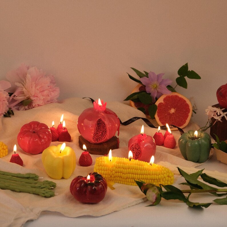 Op de afbeelding: Een verzameling kaarsen in de vorm van groenten en fruit, waaronder een rode granaatappel, gele en groene paprika, een ma&iuml;skolf en kleine aardbeien. Verschillende kaarsen branden en werpen een warme gloed op de lichte stoffen ondergrond.