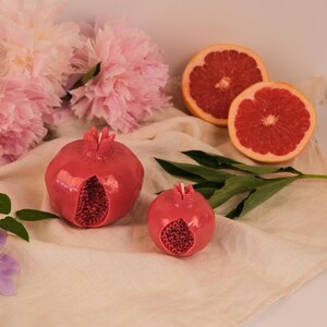Op de afbeelding: Twee granaatappelvormige kaarsen met een glanzende rode afwerking, elk met een textuur aan de binnenkant en een kleine witte lont aan de bovenkant. De kaarsen staan naast twee helften van een verse grapefruit en zachte roze pioenrozen.