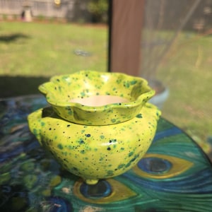 May include: A two-piece ceramic container set. The larger, rounded base has three small feet and is topped with a smaller, scalloped-edge bowl. Both pieces are a speckled, bright yellow-green with dark blue-green spots. The set is likely for decorative or functional use.