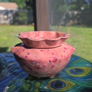 May include: A pink ceramic bowl with a scalloped rim and a speckled pattern featuring dots of red, purple, and white. The bowl has a rounded body, a wide opening, and a smooth, glossy finish, showcasing a multi-colored design on its exterior surface.