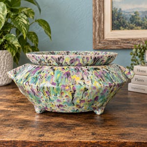 May include: A ceramic planter with a wide, shallow bowl and a larger, rounded base featuring three small feet. The surface is decorated with a marbled, multi-colored pattern in shades of teal, purple, yellow, and black, creating a vibrant effect.