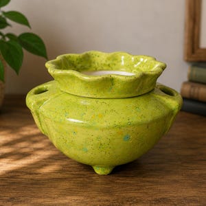 May include: A lime green ceramic flower frog with a speckled finish and a scalloped top rim. The vessel features a wide, rounded body with three circular openings on the shoulders and a small central opening, supported by three short, sturdy feet.