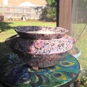 May include: A speckled pink ceramic bowl with a matching lid, resting on a peacock-themed table. The bowl has a rounded shape with small feet and a flared rim. The lid is a smaller, shallow dish. The background is a blurred outdoor scene.