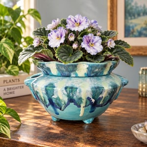 May include: A potted African violet plant with variegated white and purple petals and dark green fuzzy leaves, housed in a two-piece ceramic self-watering planter featuring a glossy turquoise, blue, and cream drip glaze finish on a base.