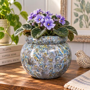 May include: A round ceramic planter with a marbled glaze in shades of blue, yellow, and grey, containing a blooming African violet plant with dark green, fuzzy leaves and purple flowers with bright yellow centers, set on a wooden surface.