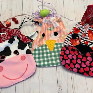 May include: Three fabric drawstring bags with unique designs. One is a cow face, another a chicken, and the third features hearts. Each bag holds a red chocolate bar and decorative accents, perfect for gifting.