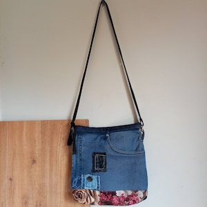 Puede incluir: Un bolso bandolera de mezclilla azul con un estampado floral en la parte inferior. El bolso tiene un bolsillo grande en la parte delantera y una correa ajustable negra.