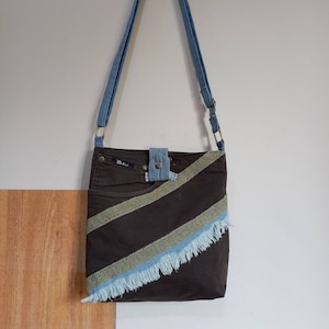 Puede incluir: Un bolso bandolera de lona marrón con una correa de mezclilla azul y detalles de flecos. El bolso tiene un ribete de tela marrón claro y un pequeño bolsillo de mezclilla azul en la parte delantera.