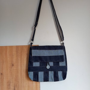 Puede incluir: Un bolso bandolera de mezclilla azul con cierre de solapa y un solo botón. El bolso tiene un patrón a rayas y una correa larga ajustable.