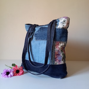 May include: A blue denim tote bag with a floral fabric patch and a dark blue denim bottom. The bag has two long straps.