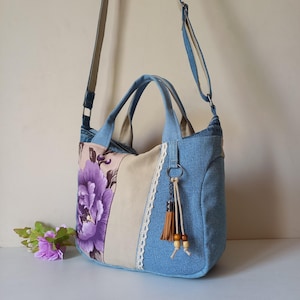 Puede incluir: Bolso tote de mezclilla con estampado floral y panel beige. El bolso presenta un diseño de peonía morada, un ribete de encaje blanco y una borla marrón. Tiene una correa para el hombro y dos asas.