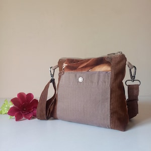May include: A brown and tan crossbody bag with a zippered top and adjustable strap. The bag features a front pocket with a decorative button and a floral print on the top panel. The bag is made of a durable fabric.